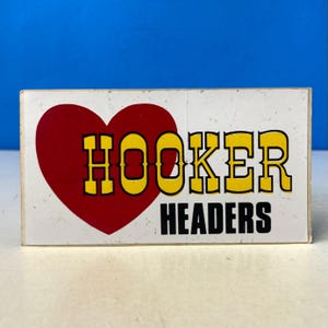 以下が含まれることがあります： 赤いハートと、黒い縁取りのある黄色の文字で「HOOKER」と書かれた長方形の看板。 「HOOKER」の下には、黒い文字で「HEADERS」と書かれています。