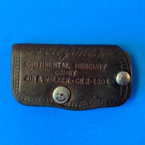 May include: A brown leather key chain with the text "Neal Jones Continental & Mercury Co. 440 S Walker Ch 2-1201" embossed on it. The key chain has two silver metal snaps.