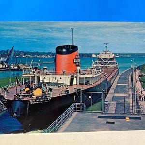 May include: Vintage postcard featuring the Tom M. Girdler ship in a canal. The ship is black with a red and black smokestack. The postcard has a blue sky and water in the background. Text on the ship reads "Tom M. Girdler Wilmington Del."