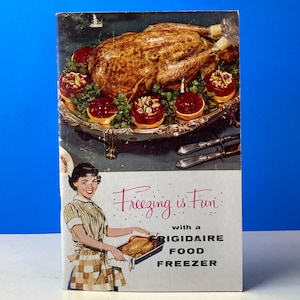 Pode incluir: Uma capa de livro de cozinha vintage com um peru assado em uma bandeja de prata com uma mulher segurando uma assadeira. O texto na capa diz "Freezing is Fun with a Frigidaire Food Freezer."