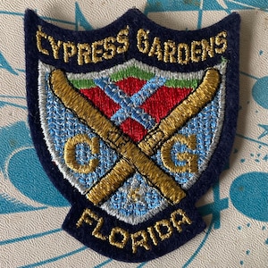 May include: A shield-shaped embroidered patch with a dark blue border. The text CYPRESS GARDENS is at the top and FLORIDA is at the bottom in gold thread. The center features two crossed gold water skis over a red and light blue background with CG.
