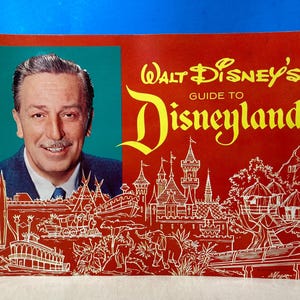 May include: A red and yellow vintage guidebook to Disneyland featuring a portrait of Walt Disney on the cover. The cover also includes a drawing of the Disneyland castle and other attractions.