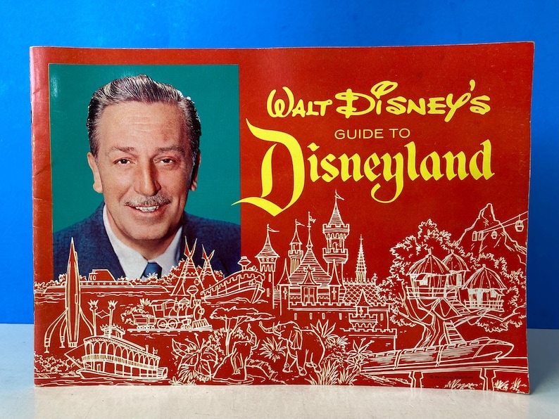 May include: A red and yellow vintage guidebook to Disneyland featuring a portrait of Walt Disney on the cover. The cover also includes a drawing of the Disneyland castle and other attractions.