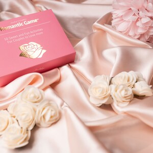 Puede incluir: Una caja rosa con letras doradas que dice "Romantic Game" y detalles de actividades para parejas, sobre una superficie de sat&eacute;n. Tambi&eacute;n hay rosas blancas y una flor rosa.