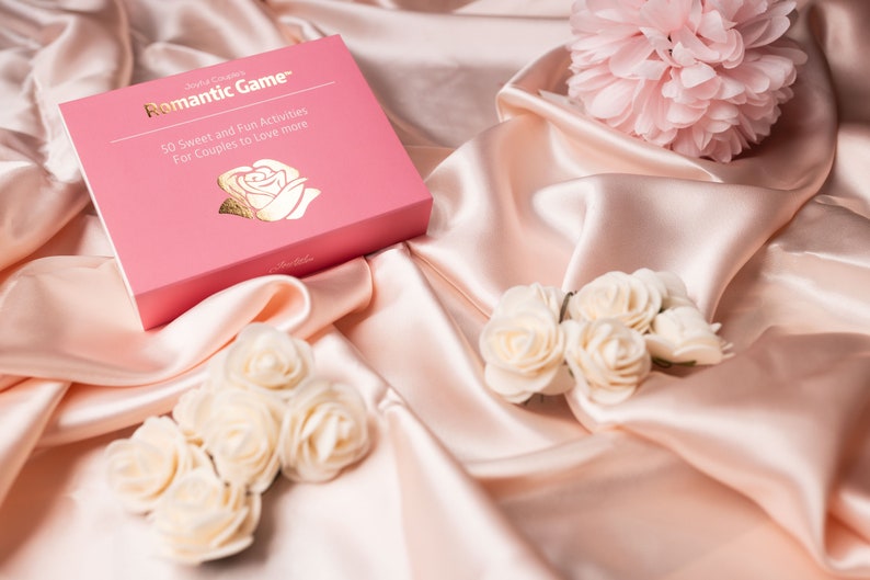 Puede incluir: Una caja rosa con letras doradas que dice "Romantic Game" y detalles de actividades para parejas, sobre una superficie de sat&eacute;n. Tambi&eacute;n hay rosas blancas y una flor rosa.