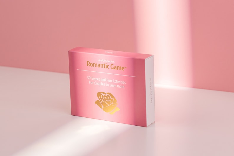 Puede incluir: Una caja rosa con las palabras "Romantic Game" en letras doradas. La caja tambi&eacute;n presenta un gr&aacute;fico de rosa dorada y el texto "50 Actividades Dulces y Divertidas Para Parejas Para Amar M&aacute;s."