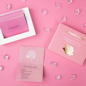 Puede incluir: Un juego rom&aacute;ntico rosa con un marco blanco que contiene tarjetas de conversaci&oacute;n rosas. La caja del juego dice "Romantic Game" con letras doradas y un gr&aacute;fico de rosa. Cristales transparentes dispersos sobre un fondo rosa.