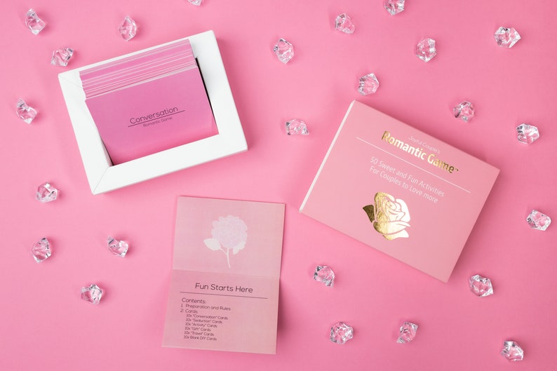 Puede incluir: Un juego rom&aacute;ntico rosa con un marco blanco que contiene tarjetas de conversaci&oacute;n rosas. La caja del juego dice "Romantic Game" con letras doradas y un gr&aacute;fico de rosa. Cristales transparentes dispersos sobre un fondo rosa.