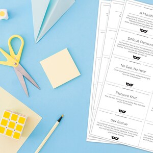 May include: A flat lay featuring stationery and printed cards on a light blue surface. Items include yellow scissors, a keyboard, a Rubik's cube, and a ruler. The cards have text and a mask graphic. The text includes "A Mouthful", "Tied Up", and "Difficult Pleasure".