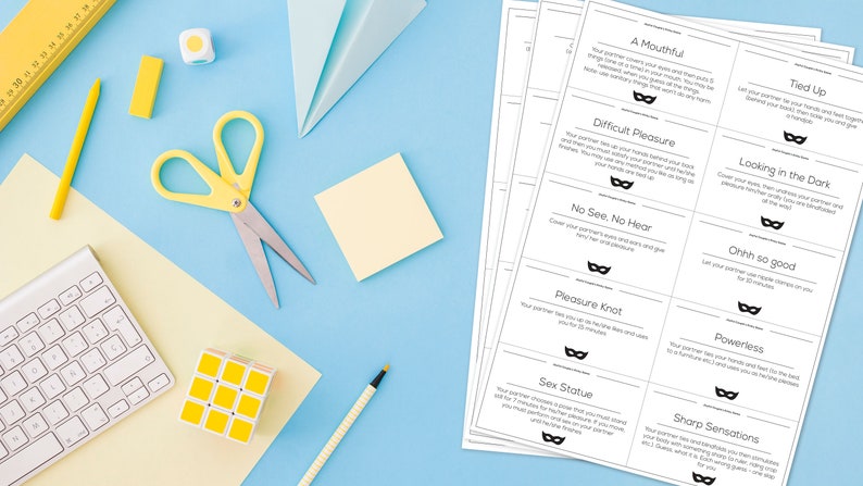 May include: A flat lay featuring stationery and printed cards on a light blue surface. Items include yellow scissors, a keyboard, a Rubik's cube, and a ruler. The cards have text and a mask graphic. The text includes "A Mouthful", "Tied Up", and "Difficult Pleasure".