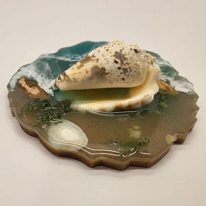 May include: A decorative resin coaster featuring a large seashell resting on a sandy beach scene. The coaster has a wave design with shades of blue and white, and the seashell is beige with brown spots.