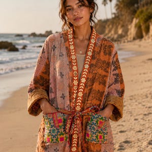 May include: A long-sleeved open-front jacket featuring a patchwork design in shades of orange, pink, and brown. The garment includes a decorative floral embroidered trim along the front edges, a matching tie belt, and two front pockets with colorful patterns.