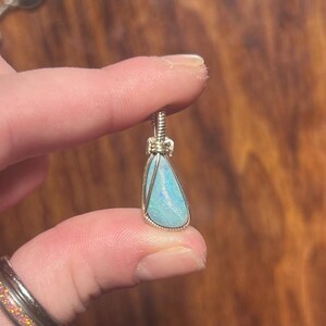 May include: A teardrop-shaped pendant featuring a light blue opal with iridescent flecks, encased in a silver-toned wire-wrapped setting with a coiled bail at the top, showcasing a smooth, polished surface and vibrant color play.
