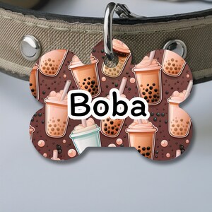 May include: A brown bone-shaped dog tag with a white background and a repeating pattern of cartoon boba tea cups. The text "Boba" is printed in black letters on the tag.
