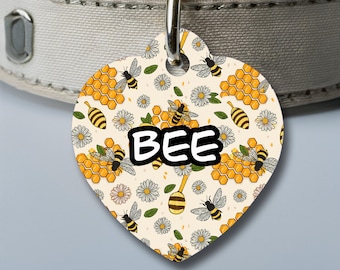 Puede incluir: Una etiqueta para perro en forma de corazón con fondo blanco y un patrón de abejas, panales y flores. La palabra "BEE" está impresa en letras negras en la etiqueta.