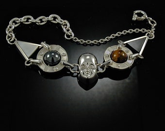 Pode incluir: Uma pulseira de prata com um fecho de caveira, um triângulo de ônix preto, uma pedra de olho de tigre e uma pedra de hematita.