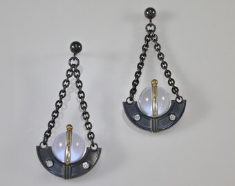 May include: A pair of silver earrings with a black finish. Each earring features a clear crystal sphere with a gold accent and two small diamonds. The sphere is suspended from a black chain.