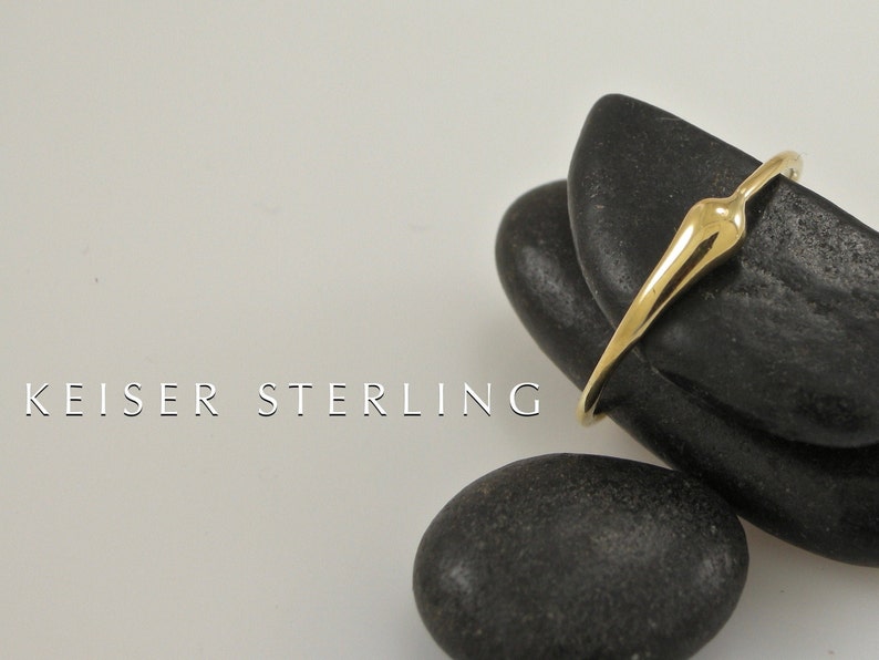 Puede incluir: Un anillo de oro con un dise&ntilde;o abstracto &uacute;nico. El anillo est&aacute; apoyado sobre dos piedras lisas negras. KEISER STERLING est&aacute; escrito en blanco debajo del anillo.