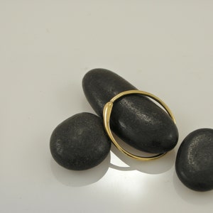 Puede incluir: Un anillo de oro con una superficie lisa y redondeada descansa sobre tres piedras negras. El anillo est&aacute; colocado sobre la piedra superior, creando una composici&oacute;n simple y elegante.