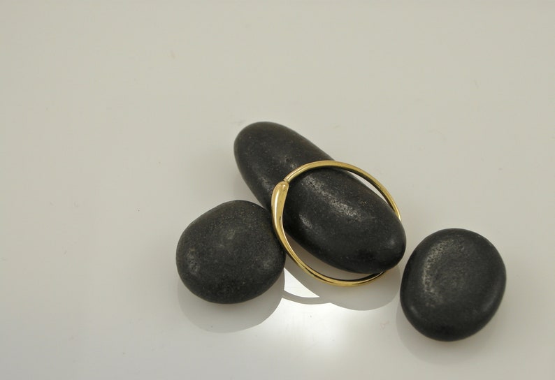 Puede incluir: Un anillo de oro con una superficie lisa y redondeada descansa sobre tres piedras negras. El anillo est&aacute; colocado sobre la piedra superior, creando una composici&oacute;n simple y elegante.