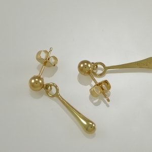 Pode incluir: Um par de brincos de ouro com uma pequena bola no topo e um pingente longo, fino e em forma de l&aacute;grima.