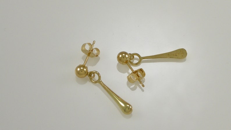 Pode incluir: Um par de brincos de ouro com uma pequena bola no topo e um pingente longo, fino e em forma de l&aacute;grima.