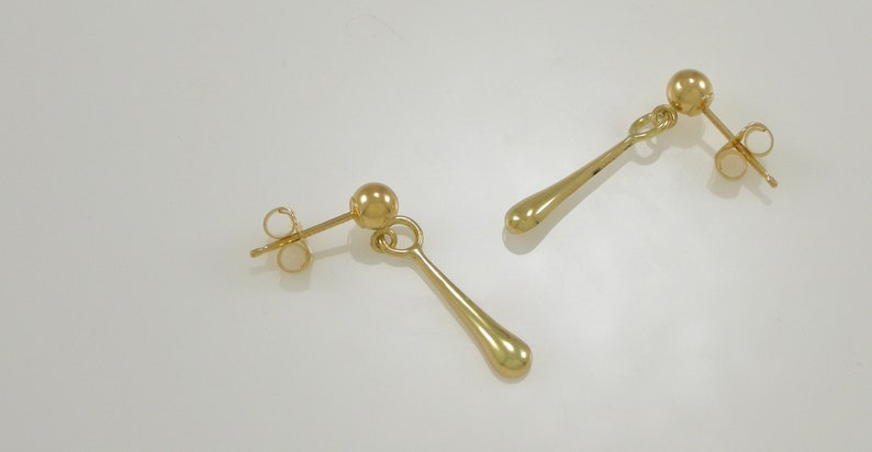 Pode incluir: Um par de brincos de gota de ouro com uma pequena bola no topo e um fecho de poste.
