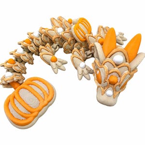 Könnte beinhalten: Eine bewegliche Drachenfigur mit cremefarbenem Körper, orangefarbenen Akzenten und weißen kugelförmigen Details. Der Drache hat einen segmentierten Schwanz, der in einem kürbisförmigen Teil endet, orangefarbene Hörner und eine strukturierte Oberfläche.