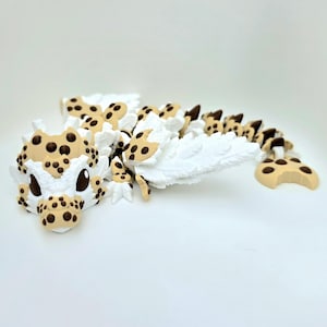 May include: A multi-jointed articulated dragon figure featuring a white body with tan accents and dark brown spots. The creature has large dark eyes, textured wings, and a segmented tail that curves along the surface, resembling a cookie design.