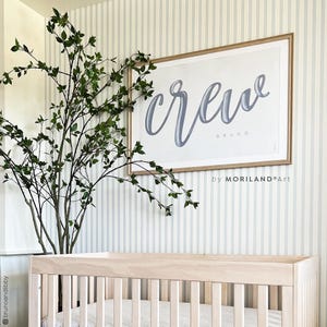 Op de afbeelding: Een ingelijste print met het woord "crew" in een cursief lettertype, met de naam "Bruno" eronder. De print zit in een licht houten frame en hangt aan een gestreepte muur. Een wieg en bladerrijke takken staan op de voorgrond.