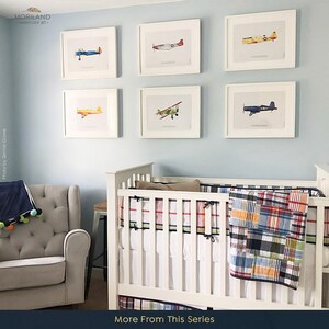 May include: Six framed watercolor paintings of vintage airplanes in a nursery setting. The paintings are hung on a light blue wall above a white crib with a plaid quilt. The crib is next to a gray armchair.