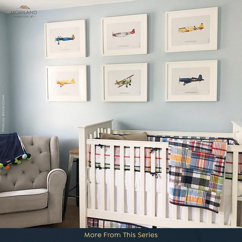 May include: Six framed watercolor paintings of vintage airplanes in a nursery setting. The paintings are hung on a light blue wall above a white crib with a plaid quilt. The crib is next to a gray armchair.