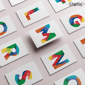 May include: A set of colorful, geometric alphabet letter cards. Each card features a different letter of the alphabet, made up of colorful, geometric shapes. The cards are arranged in a grid pattern on a light pink surface. The text "The Original MORILAND Art" is visible in the top right corner of the image.