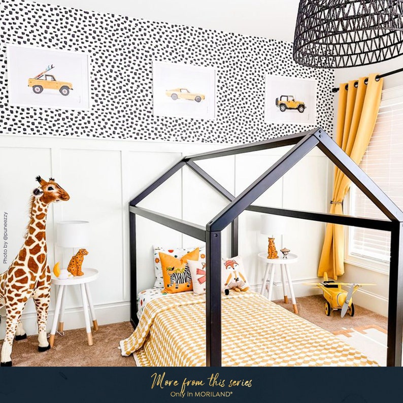 May include: A black wooden house-shaped bed frame with a yellow and white checkered comforter. The bed is decorated with pillows featuring animal prints and a stuffed giraffe stands next to the bed. The wall behind the bed is decorated with a black and white polka dot pattern and three framed prints of yellow toy cars.