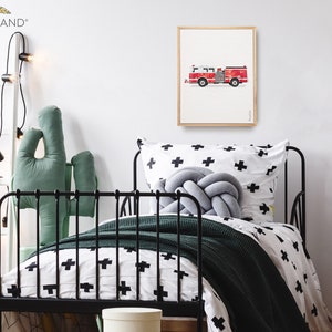 Op de afbeelding: Een ingelijste print van een rode brandweerwagen op een witte achtergrond. De print hangt aan een witte muur in een kinderkamer. De slaapkamer heeft een zwart metalen bed met een wit en zwart gestreept dekbedovertrek, een groen cactus kussen en een wit nachtkastje met een groene cactusvormige container.