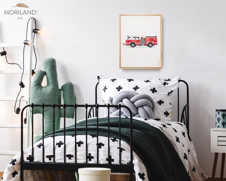 Op de afbeelding: Een ingelijste print van een rode brandweerwagen op een witte achtergrond. De print hangt aan een witte muur in een kinderkamer. De slaapkamer heeft een zwart metalen bed met een wit en zwart gestreept dekbedovertrek, een groen cactus kussen en een wit nachtkastje met een groene cactusvormige container.