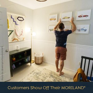 May include: A person hangs framed prints of vintage cars on a white wall. The prints are in various colors, including red, yellow, blue, and green. The prints are all in the same style, with a white background and black outlines. The person is wearing a blue shirt and brown shorts. The room is decorated in a modern style, with a white rug and a black bookshelf.