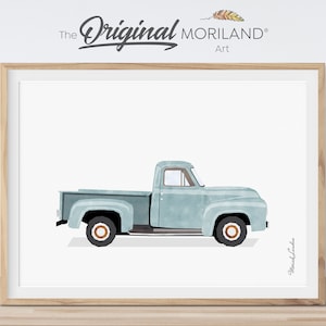 May include: A framed print of a light blue vintage pickup truck illustration. The truck has black tyres with brown rims, set against a white background. The artwork is signed and includes the text "The Original Moriland Art".