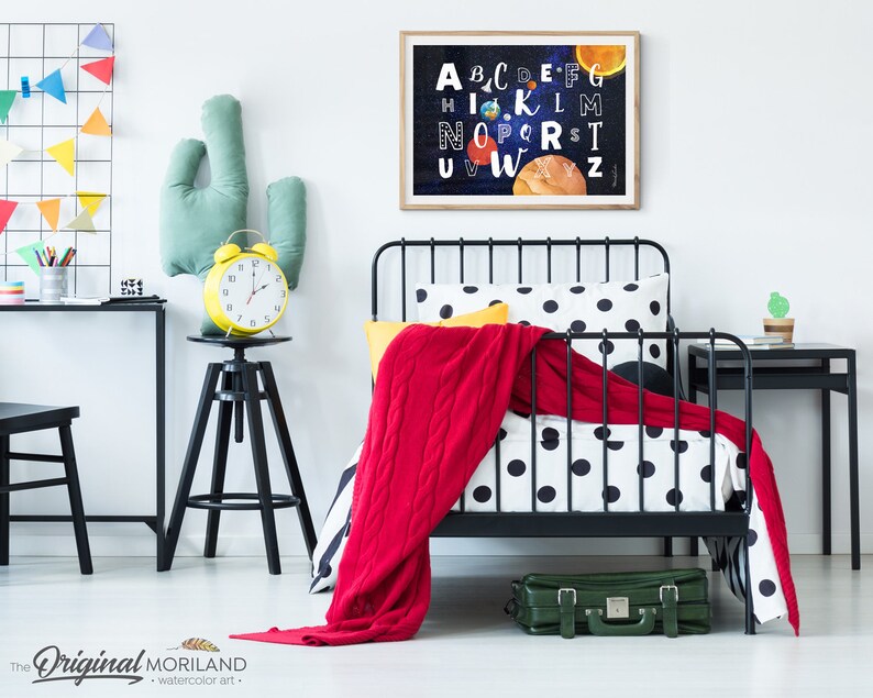 May include: A black metal bed frame with a white and black polka dot duvet cover and a red blanket. A yellow alarm clock sits on a black stool next to a green cactus pillow. A framed print of the alphabet with planets and stars hangs on the wall.