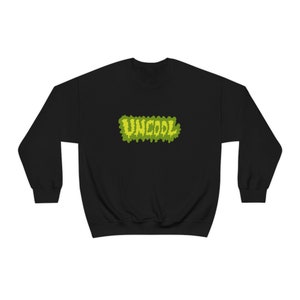Puede incluir: Sudadera negra con un gráfico verde y amarillo que dice "UNCOOL" en una fuente que gotea.
