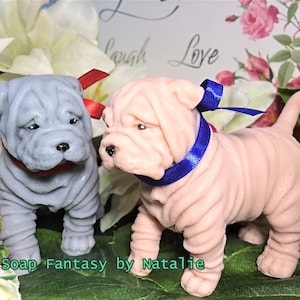 May include: Two handmade soap sculptures of Shar-Pei puppies. One puppy is gray with a red ribbon around its neck. The other puppy is pink with a blue ribbon around its neck. The soap sculptures are on a green leafy background. Soap Fantasy by Natalie.