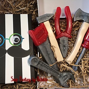 May include: A gift box filled with brown shredded paper and five toy tools made of soap. The tools are a red saw, a red screwdriver, a silver wrench, a silver hammer, and a pair of red pliers. The box has a black and white striped lid with a cartoon face on it. The face has googly eyes, a black mustache, and the text "Soap Fantasy by Natalie".