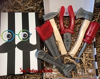 Op de afbeelding: Een cadeaudoos gevuld met bruin, versnipperd papier en vijf speelgoedgereedschappen van zeep. De gereedschappen zijn een rode zaag, een rode schroevendraaier, een zilveren moersleutel, een zilveren hamer en een rode tang. De doos heeft een zwart-wit gestreept deksel met een cartoon gezicht erop. Het gezicht heeft knikkerogen, een zwarte snor en de tekst "Soap Fantasy by Natalie".