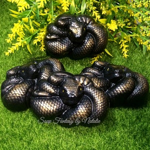 May include: Four black and gold soap bars shaped like coiled snakes with yellow eyes. The soap bars are on a bed of green grass.