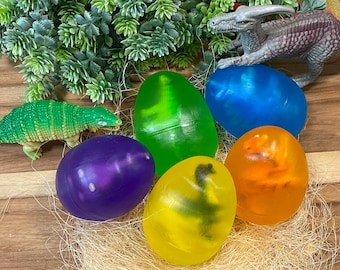 Op de afbeelding: Zes doorzichtige plastic eieren met dinosaurusfiguurtjes erin. De eieren zijn in verschillende kleuren, waaronder paars, groen, blauw, geel en oranje. De eieren liggen op een bedje van stro.