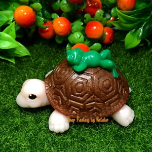 May include: A brown turtle with a green frog sleeping on its back. The turtle has a detailed shell and white feet. The turtle and frog are on a green grassy surface.