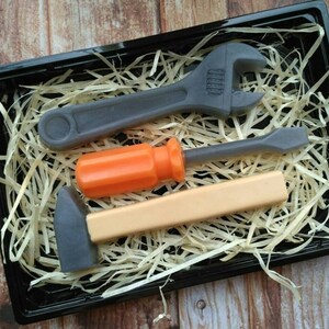 Op de afbeelding: Een set van drie speelgoedgereedschappen in een zwarte bak met gescheurd papier. De gereedschappen zijn een moersleutel, een schroevendraaier en een hamer. De moersleutel en de hamer zijn grijs, en de schroevendraaier heeft een oranje handvat.