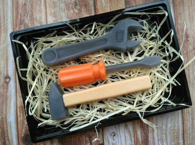 Op de afbeelding: Een set van drie speelgoedgereedschappen in een zwarte bak met gescheurd papier. De gereedschappen zijn een moersleutel, een schroevendraaier en een hamer. De moersleutel en de hamer zijn grijs, en de schroevendraaier heeft een oranje handvat.