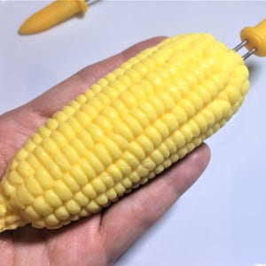 以下が含まれることがあります： 黄色いトウモロコシの穂先と黄色いプラスチック製の串。トウモロコシは手で持たれている。