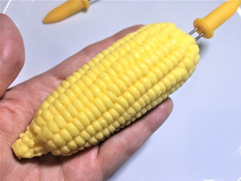 以下が含まれることがあります： 黄色いトウモロコシの穂先と黄色いプラスチック製の串。トウモロコシは手で持たれている。